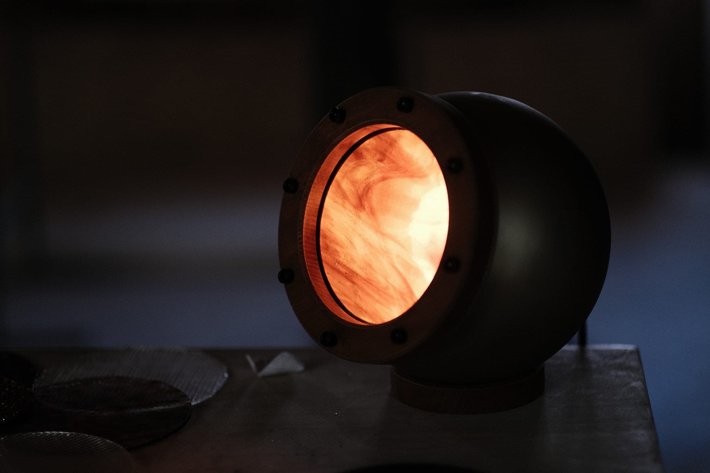 Lampe Abysse allumée, vue de trois quarts : sphère d’acier, anneau de bois et verre marbré orange